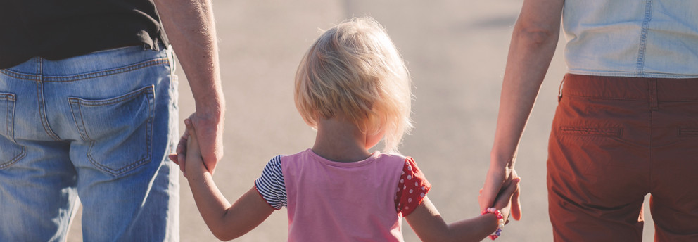 Psychologische-Pädagogische Ambulanz Rückansicht eines blonden Kindes, welches zwischen seinen Eltern läuft, ein Elternteil jeweils an einer Hand.